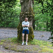 Man standing in forest with arms crossed wearing grey tshirt and navy blue lounge short.