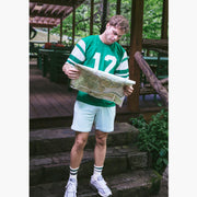 Man standing on stone steps wearing green/white jersey with number 12 holding a map and also wearing white lounge shorts.