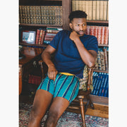 Man sitting in library wearing green/blue/yellow stripe slim fit boxers and blue shirt.